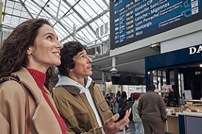 Foto de un hombre y una mujer en una estación delante de una pantalla que muestra las salidas y llegadas de trenes.